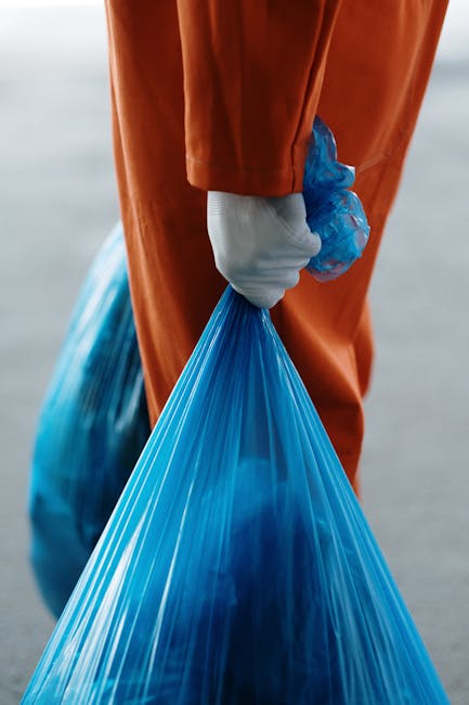 A person wearing orange overalls and a white glove is holding a large, thick, blue plastic rubbish bag tightly by the gathered top, with their arm positioned in the center of the image. The bag appears filled with waste material, causing it to bulge slightly in the middle. The background is blurred but suggests an outdoor setting, possibly on a pavement or driveway, with neutral lighting that highlights the glossy, semi-translucent texture of the plastic bag. The scene emphasizes the handling and disposal of bulk waste, reflective of private rubbish removal or collection services. The individual's clothing and glove indicate a hygienic, professional approach to waste management, consistent with the services offered by companies like House Clearance Edgware. This scene visually supports concepts of on-site clearance and independent waste handling, involving manual lifting and collection of rubbish as part of efficient waste removal solutions.