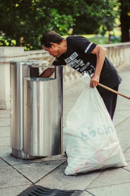 A middle-aged man with short dark hair is seen outdoors on a paved area, leaning forward as he disposes of a white bag filled with rubbish into a silver, cylindrical metal waste bin. The bin has a shiny, reflective surface and an open top designed for easy emptying. The man is dressed in a dark t-shirt with white text and graphics on the front, and appears to be focused on placing the bag into the bin, which is part of a public waste disposal station. In his right hand, he holds the white rubbish bag, while his left hand is resting on the edge of the bin. The background features a low stone wall and lush green foliage, indicating a park or garden setting with ample natural light. The overall scene reflects a typical situation involving private or independent waste disposal outdoors, and may relate to rubbish removal services that support on-site clearance and waste collection outside of municipal systems, aligning with the context of avoiding hidden fees for rubbish removal in Edgware, Edgware.