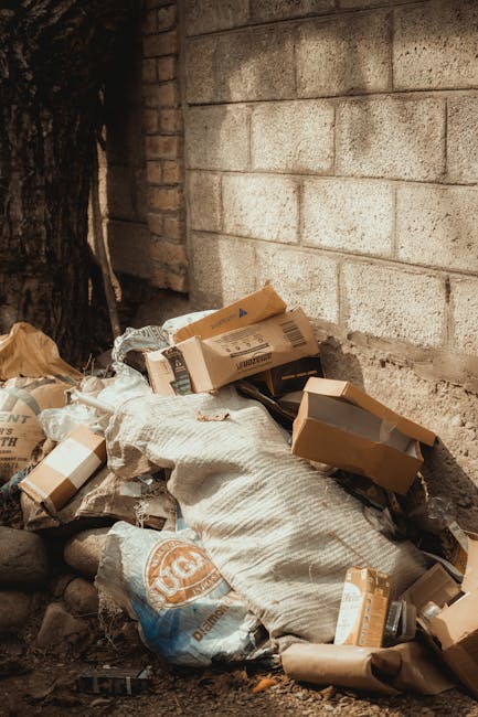 A person wearing orange overalls and a white glove is holding a large, thick, blue plastic rubbish bag tightly by the gathered top, with their arm positioned in the center of the image. The bag appears filled with waste material, causing it to bulge slightly in the middle. The background is blurred but suggests an outdoor setting, possibly on a pavement or driveway, with neutral lighting that highlights the glossy, semi-translucent texture of the plastic bag. The scene emphasizes the handling and disposal of bulk waste, reflective of private rubbish removal or collection services. The individual's clothing and glove indicate a hygienic, professional approach to waste management, consistent with the services offered by companies like House Clearance Edgware. This scene visually supports concepts of on-site clearance and independent waste handling, involving manual lifting and collection of rubbish as part of efficient waste removal solutions.