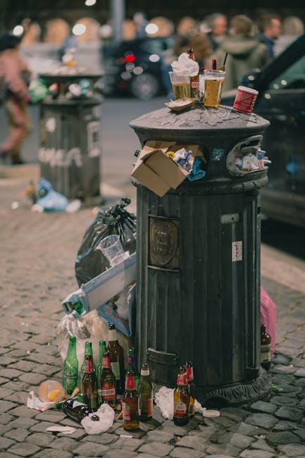 The image depicts an outdoor scene with an extensive collection of waste materials arranged for disposal. Several large stacks of cardboard boxes, primarily branded with labels such as 'fresh fruits,' are neatly piled, indicating collection of produce packaging. The boxes are mostly flattened, with some still partially assembled, showing bright and colorful printed designs on a glossy surface. To the right, metal wire cages filled with various paper and cardboard packaging are visible, with some boxes open and contents partly visible, including brown shipping cartons and smaller boxes. In the foreground, multiple plastic bins in green, red, and orange are positioned on the concrete surface, some with their lids closed, while others are open or tipped over. A few loose items, such as a blue plastic bag and a black trash bag, are scattered among the bins and packaging. The environment appears to be a residential driveway or yard, with a backdrop of greenery, trees, and distant rooftops under natural daylight, emphasizing the scene of waste collection and removal, potentially related to private disposal or rubbish services as offered by House Clearance Edgware.