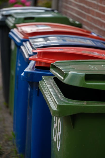 A row of large, rectangular wheelie bins lined up against a brick wall on a residential pavement. The bins are made of durable plastic with textured surfaces and are in various colours including green, blue, red, and dark green, each topped with a matching lid that is closed. They are positioned side by side, with the green and blue bins in the foreground and the red and dark green bins visible further back, creating a visual sequence of waste containers for household rubbish collection. The environment appears to be outdoors in a suburban setting, with soft natural lighting highlighting the different surface textures and contrasting colours of the bins. The scene suggests an organized approach to waste management, potentially related to private or independent rubbish collection services such as those provided by House Clearance Edgware, supporting non-local-authority rubbish disposal or on-site clearance activities.