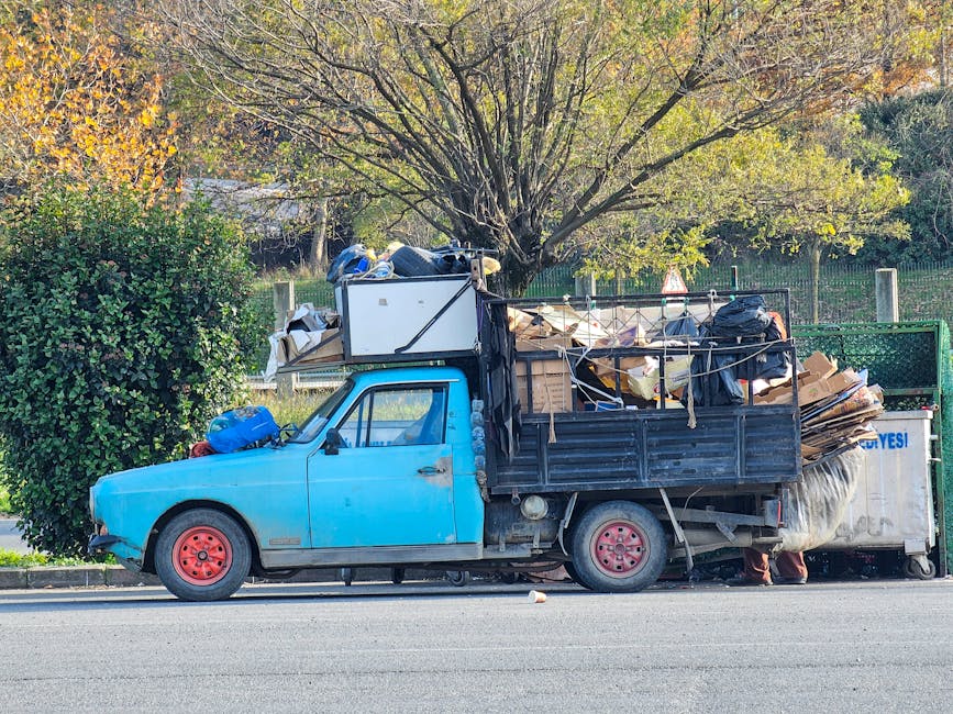 A light blue, compact flatbed truck is parked on the side of a street with its cargo area heavily loaded with assorted rubbish and discarded materials, including flattened cardboard boxes, black plastic bags, and large wooden panels. The vehicle's paintwork shows signs of wear and minor rust, with red-painted wheels matching the overall utilitarian appearance. The load extends beyond the truck's sides, secured with ropes and straps, indicating a private collection or on-site clearance activity. In the background, a tree with green and golden leaves suggests an autumn setting, and a metal fence runs parallel to the street, partially obscured by the foliage. The surrounding environment appears to be a residential or semi-urban area with natural greenery, and the scene is set in daylight with clear, unobstructed visibility, reflecting a typical rubbish removal scenario where local or private waste collection is undertaken outside of official municipal services. House Clearance Edgware specializes in alternative waste handling, as demonstrated by the heavy load prepared for disposal.