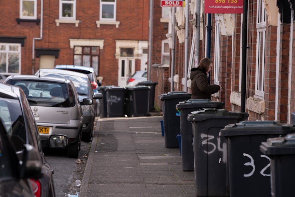 A woman standing on a sidewalk beside a row of black waste bins with gray lids, arranged along the front of a red brick residential building. The bins are labeled with white spray-painted numbers, and some have newspapers or plastic bags resting on top. Several parked cars line the street on the left side, with a mix of silver, black, and gray vehicles, including a small hatchback and a compact vehicle. The background features additional brick houses with white-framed windows, and a sign indicating property sale hangs on the building. The scene is captured during daylight, with natural lighting casting soft shadows. The pavement appears clean apart from a few scattered pieces of litter, emphasizing the context of waste disposal and rubbish collection services, such as those provided by House Clearance Edgware, which caters to private rubbish removal needs in the Broadfields Estate area.