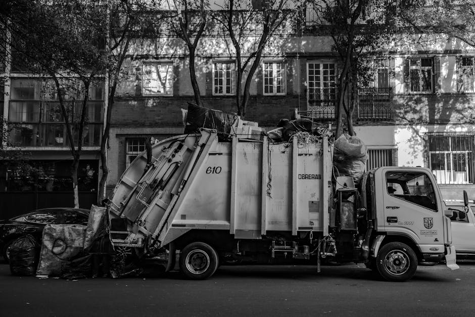 A side view of a large rubbish collection truck parked on a city street, with the truck's cab positioned on the right. The truck features a metal body with visible mechanical components, hoses, and a loading mechanism at the rear, used for waste collection and disposal. The vehicle's surface appears weathered and utilitarian, with a small identification number '610' and some text partially visible on the side. In front of the truck, several black garbage bags and bundled items are placed on the pavement, indicating ongoing waste collection and clearance activities. The background includes a multi-story residential building with large windows and a balcony, as well as leafless trees casting shadows on the building façade, hinting at an urban environment and possibly the autumn or winter season. The overall scene reflects a routine waste collection process in a residential area, highlighting the role of private rubbish removal services like House Clearance Edgware in managing household and property clearance tasks.