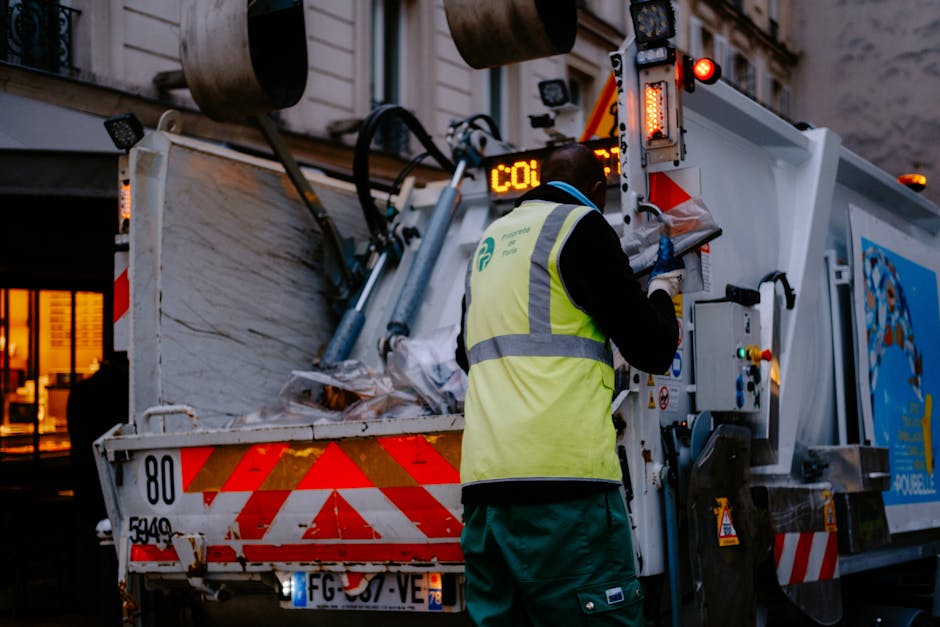 A side view of a large rubbish collection truck parked on a city street, with the truck's cab positioned on the right. The truck features a metal body with visible mechanical components, hoses, and a loading mechanism at the rear, used for waste collection and disposal. The vehicle's surface appears weathered and utilitarian, with a small identification number '610' and some text partially visible on the side. In front of the truck, several black garbage bags and bundled items are placed on the pavement, indicating ongoing waste collection and clearance activities. The background includes a multi-story residential building with large windows and a balcony, as well as leafless trees casting shadows on the building façade, hinting at an urban environment and possibly the autumn or winter season. The overall scene reflects a routine waste collection process in a residential area, highlighting the role of private rubbish removal services like House Clearance Edgware in managing household and property clearance tasks.