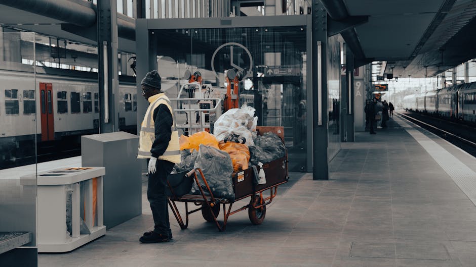 At a modern train station platform, a worker dressed in dark clothing, a high-visibility vest, and a knit cap stands next to a red-handled trolley loaded with large bags of various materials, including clear plastic, black, and orange trash bags, along with some loose items that appear to be discarded rubbish. The trolley is positioned on a wide, paved surface with a neutral grey finish, adjacent to a glass-enclosed waiting area supported by metal beams. Behind the person, there are two stationary trains with sleek, metallic exteriors, and a few passengers are seen further down the platform. The environment is well-lit, with natural daylight illuminating the scene and casting soft shadows. The context suggests an independent waste collection activity, possibly related to on-site clearance or rubbish removal services at a public transportation hub, managed by a professional waste handling company. This scene reflects typical rubbish management practices involving the collection and transportation of waste from urban locations for disposal or recycling purposes, ensuring cleanliness and order in frequented public spaces.