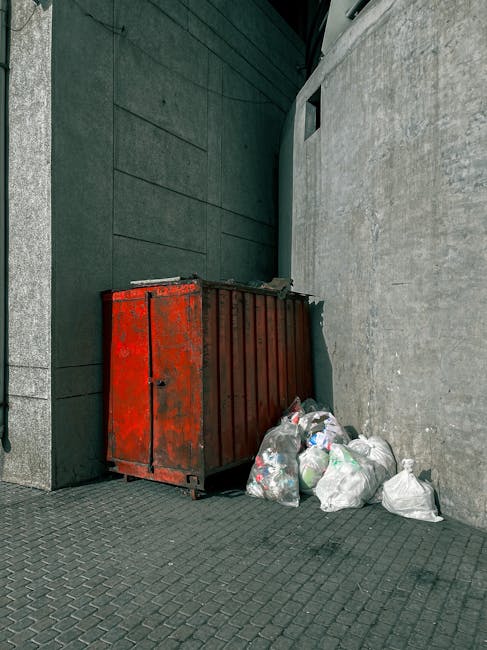 A red metal waste disposal container positioned against a dark green wall on the left and a textured light grey concrete wall on the right, situated on an outdoor paved surface with a rubber matting pattern. Several white and transparent plastic rubbish bags, filled with assorted waste, are piled next to or leaning against the container, indicating ongoing rubbish collection or disposal activity. The scene appears to be in an urban alleyway or backstreet environment, with a small, rectangular window or ventilation opening visible high up on the grey wall. The overall setting suggests an area designated for rubbish accumulation awaiting collection or private waste handling services, with natural lighting highlighting the textures of the metal, concrete, and plastic bags. House Clearance Edgware’s rubbish removal services are suited for efficiently managing waste in such functional outdoor spaces where independent collection or on-site clearance is required for timely waste management.